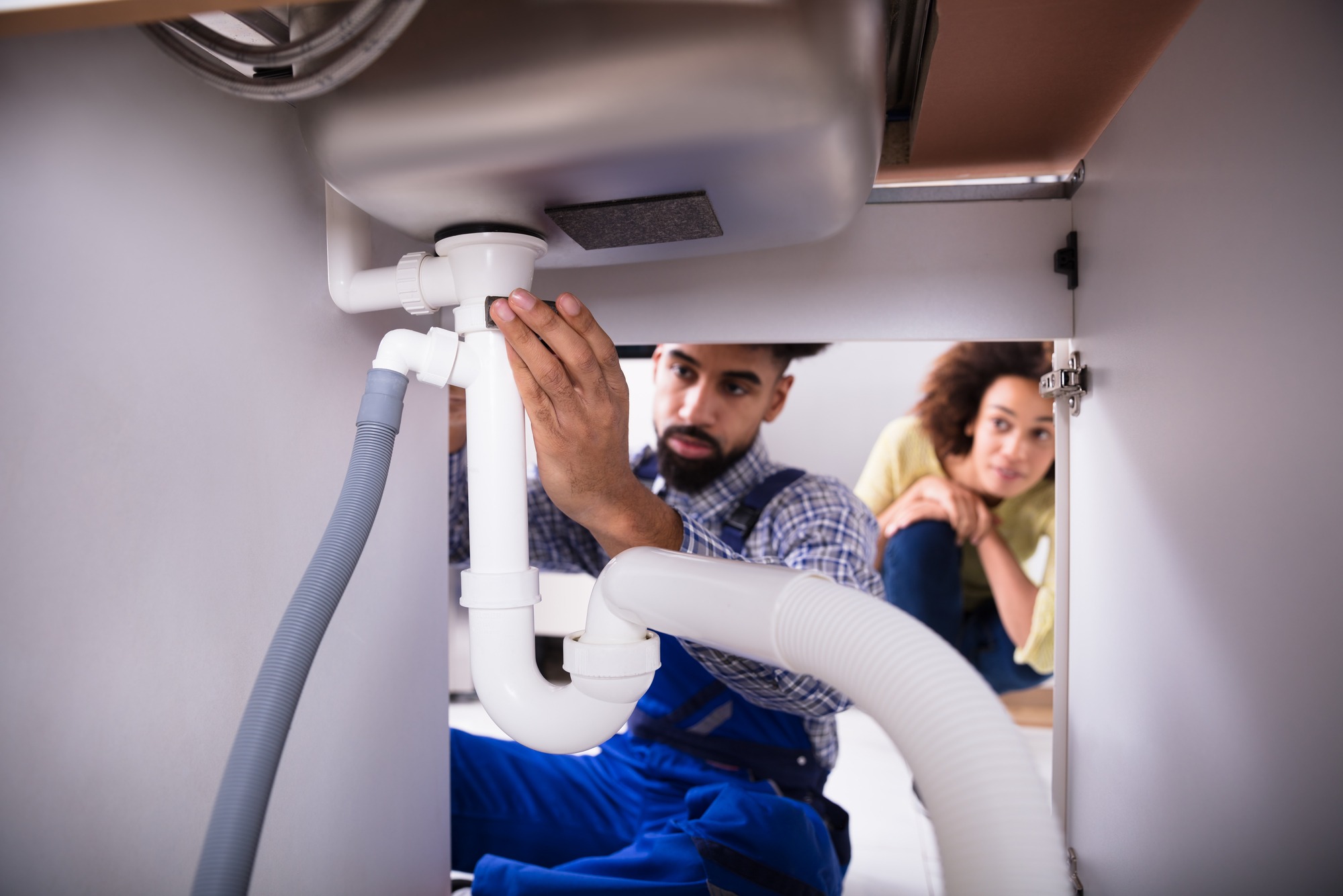 A plumber inspects and adjusts pipes under a kitchen sink, while a concerned homeowner observes. The image illustrates home plumbing maintenance.