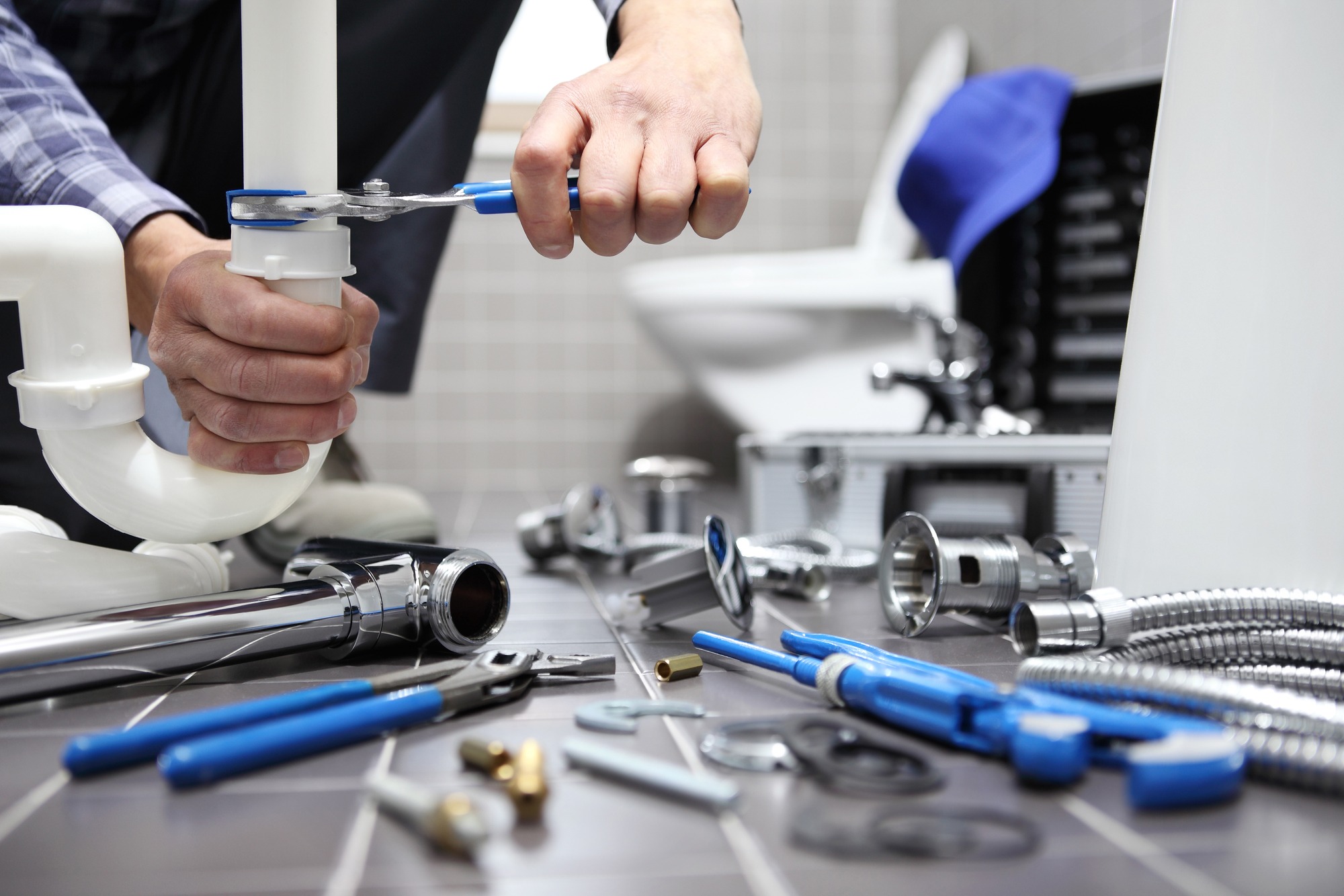 A plumber uses a wrench to tighten a pipe under a sink, surrounded by various plumbing tools and parts on the floor, showcasing a home repair scenario.