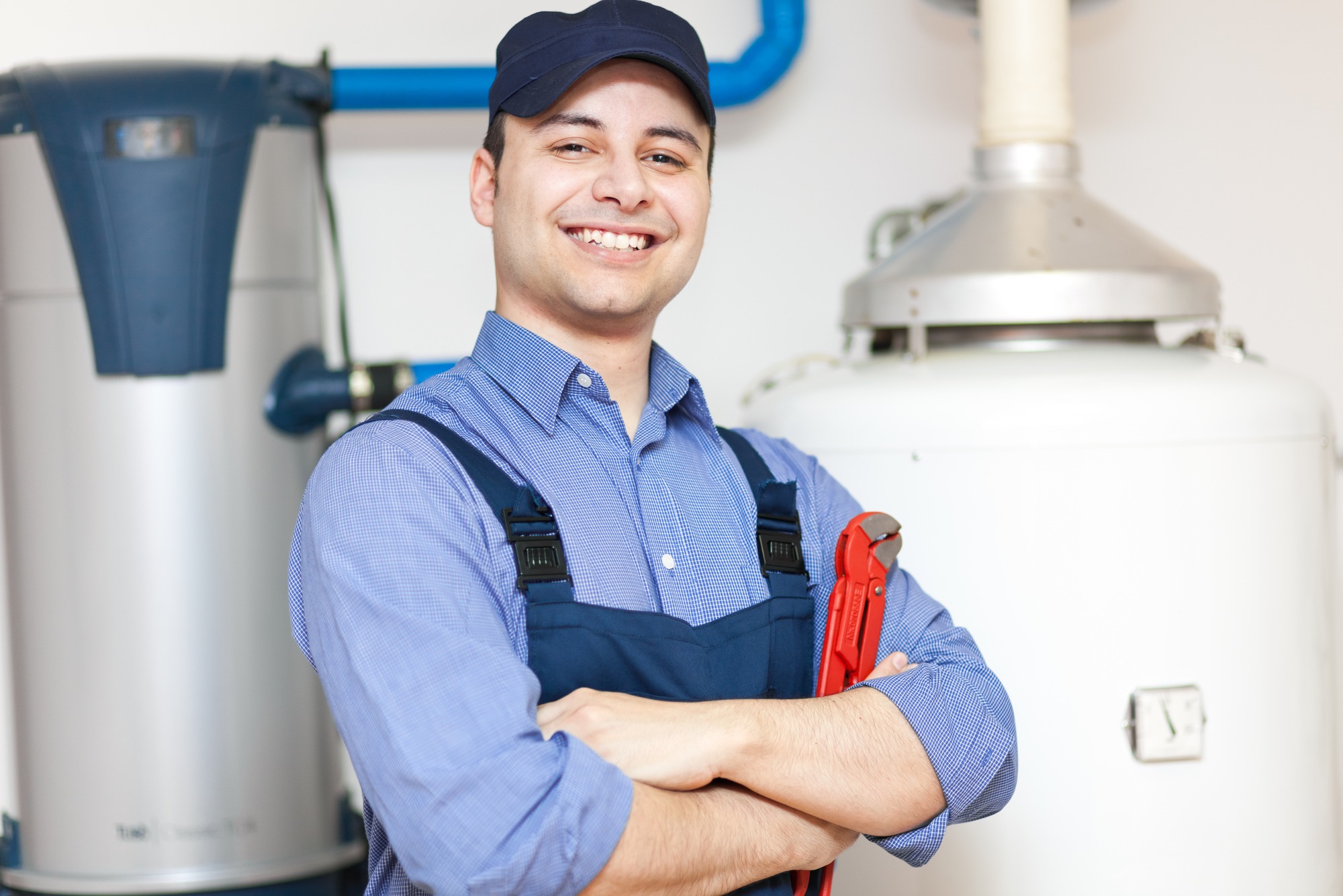 Smiling plumber in uniform stands confidently with arms crossed, holding a red wrench, in a well-lit utility room with plumbing equipment.