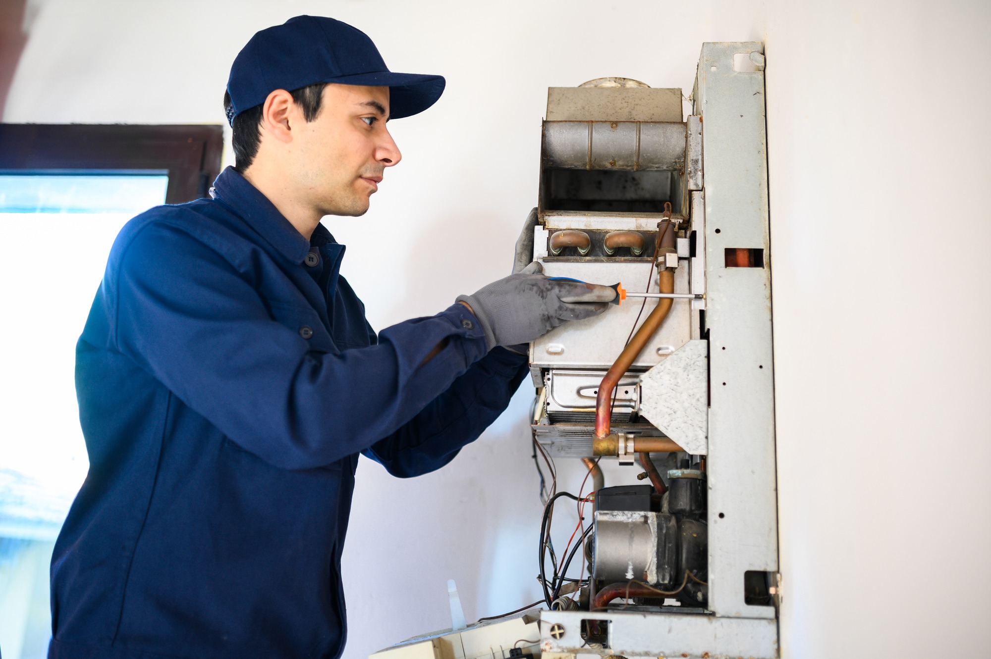 A technician in gloves adjusts a wall-mounted heating unit with a screwdriver, demonstrating routine maintenance and expertise in HVAC work.